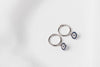 A pair of  huggies earring silver hoop adorned with a blue evil eye plate, displayed on a white background.