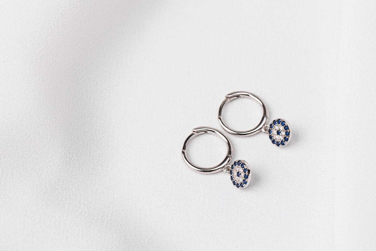 A pair of  huggies earring silver hoop adorned with a blue evil eye plate, displayed on a white background.