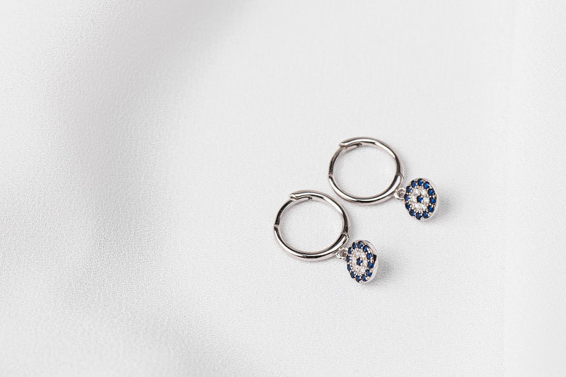 A pair of  huggies earring silver hoop adorned with a blue evil eye plate, displayed on a white background.
