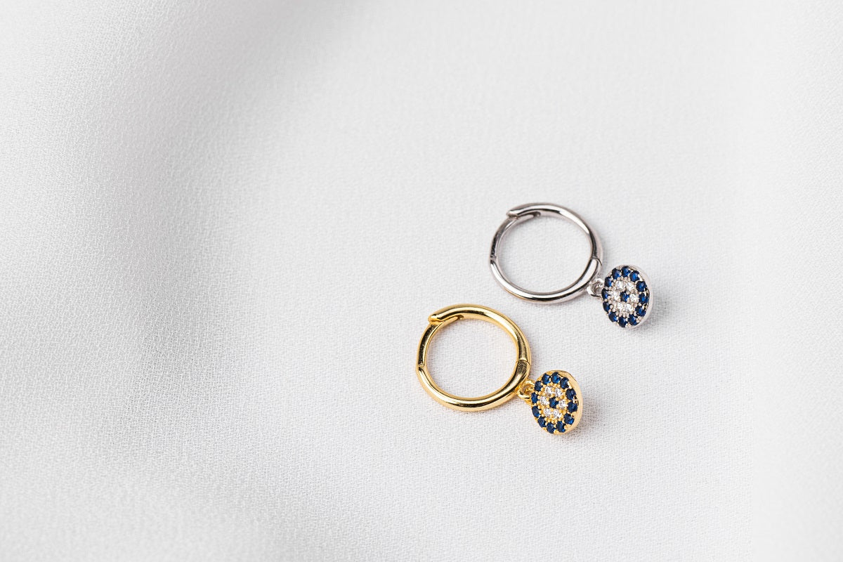Two huggies earrings with one featuring a silver hoop and the other with a gold vermeil hoop, both adorned with a blue evil eye plate, displayed on a white background.