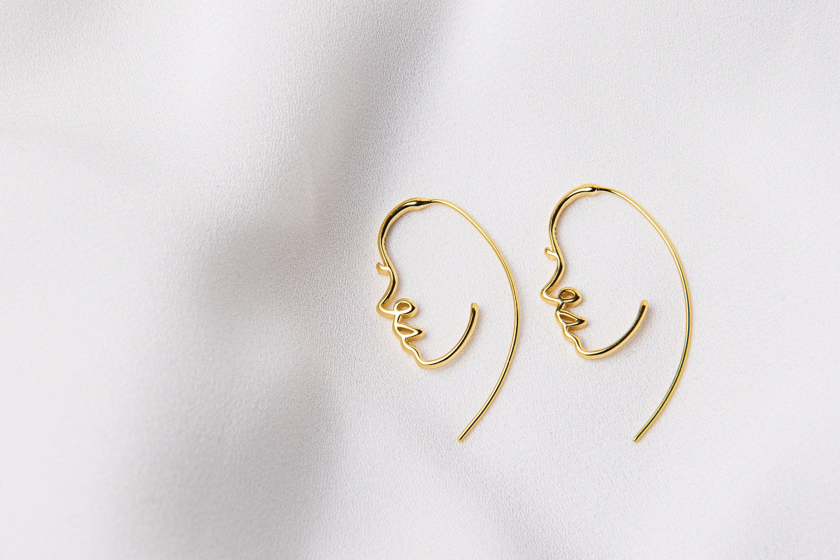 A pair of gold vermeil earrings with a minimalist design depicting a face, placed on a white fabric surface.