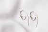 A pair of silver earrings with a minimalist design depicting a face, placed on a white fabric surface.