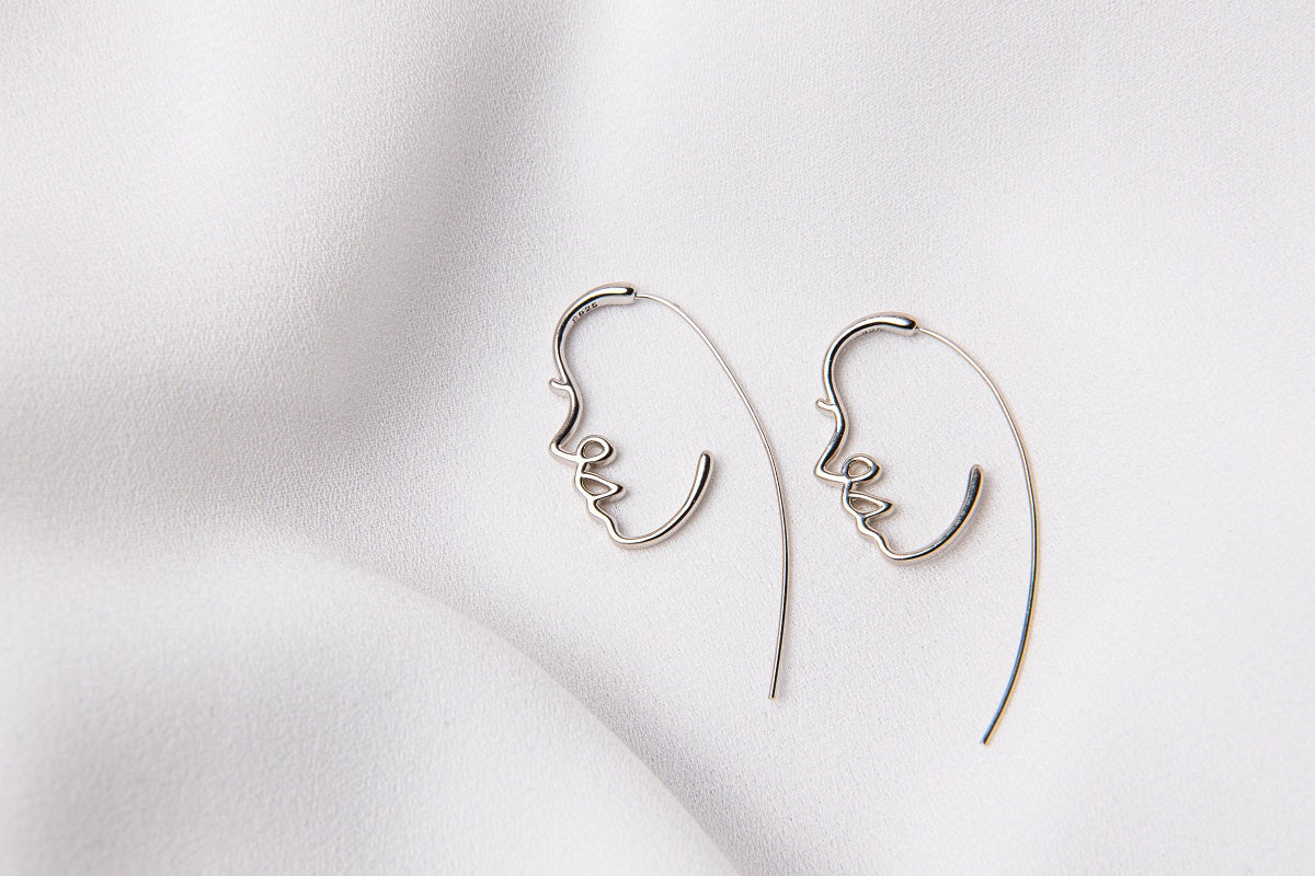 A pair of silver earrings with a minimalist design depicting a face, placed on a white fabric surface.