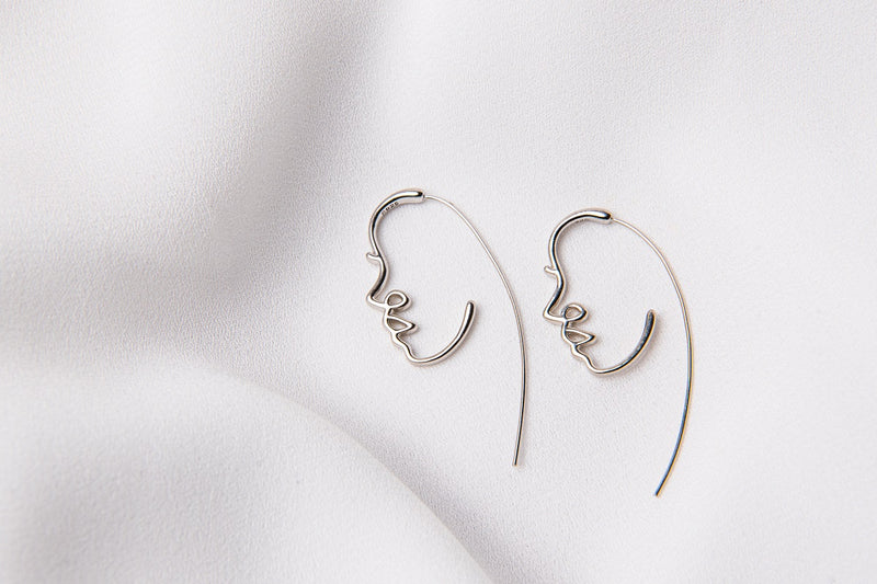 A pair of silver earrings with a minimalist design depicting a face, placed on a white fabric surface.