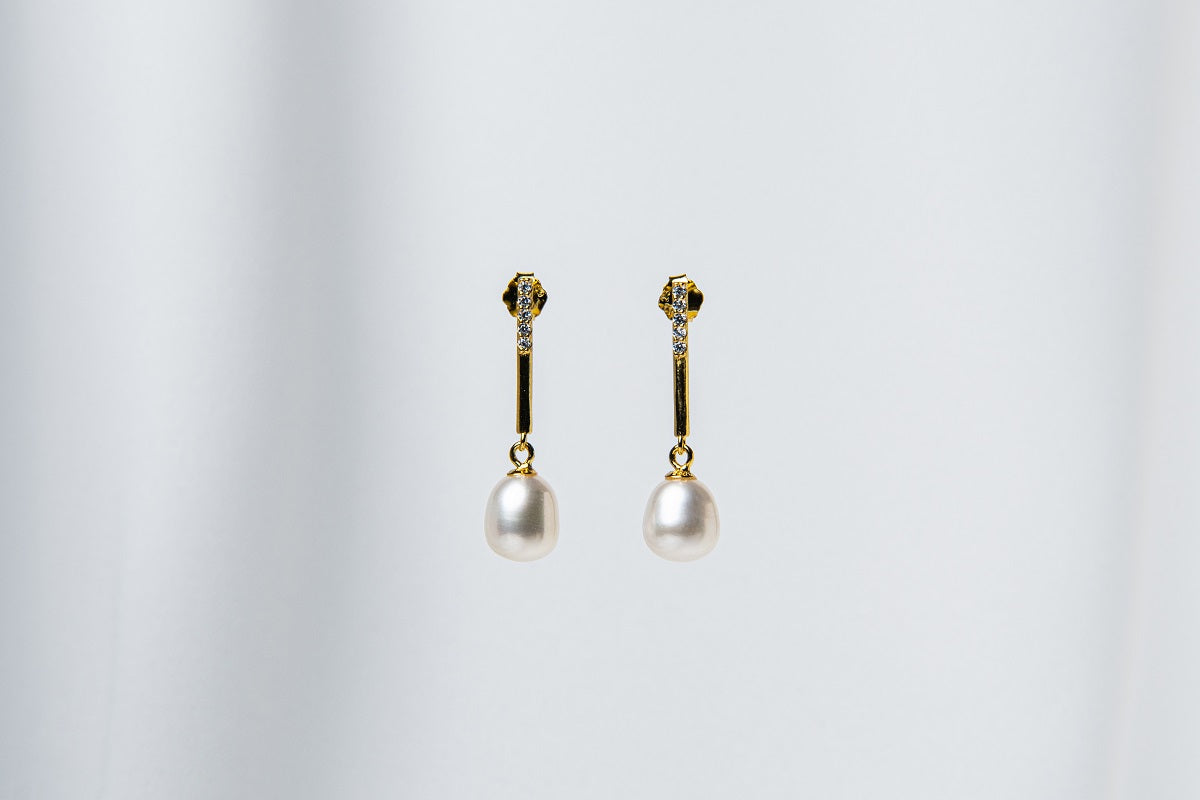 A pair of dangle earrings with a drop design, featuring pearl earrings and gold-colored metal backs, displayed against a white fabric background.