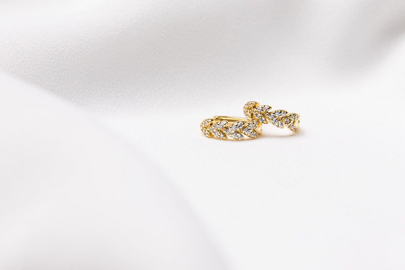 A pair of gold vermeil huggies earrings with leaf patterns with cubic zirconia stones, displayed on a white satin background.