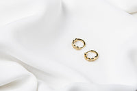 A pair of gold vermeil huggies earrings with leaf patterns with cubic zirconia stones, displayed on a white satin background.