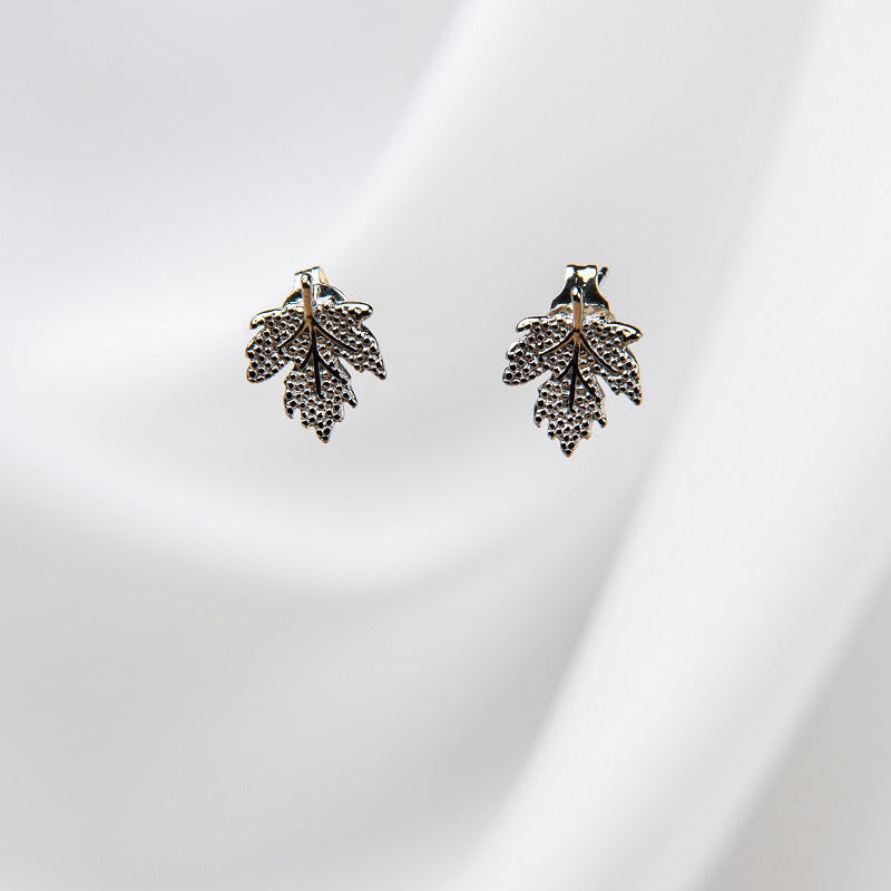 A pair of maple leaf-shaped stud earrings with a gold vermeil finish on a white silky background.