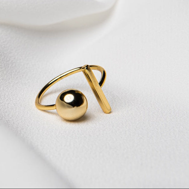 Close up of a gold vermeil ring featuring a bar and a ball on a white fabric background.