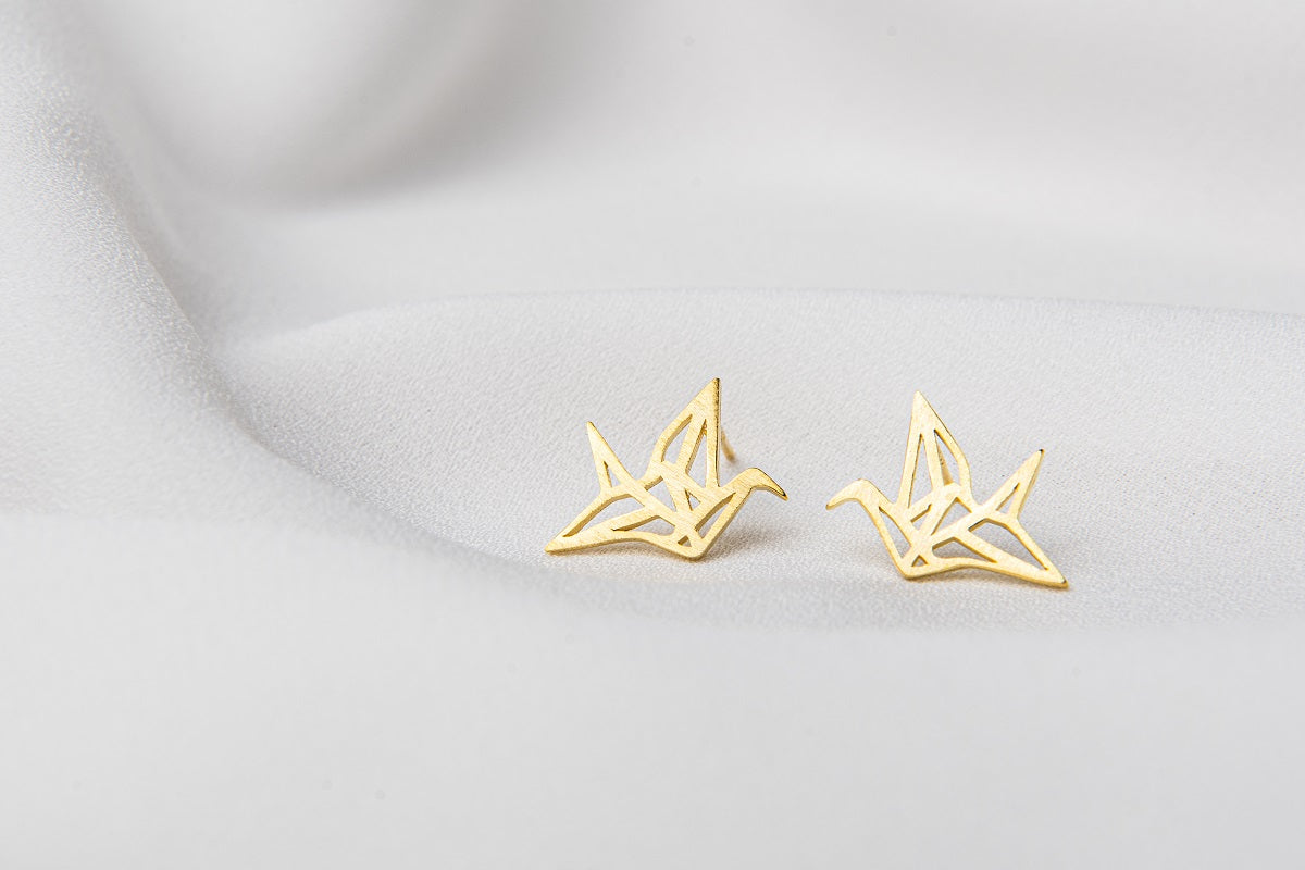 A pair of gold stud earrings designed in a origami crane shape, placed on a white surface.