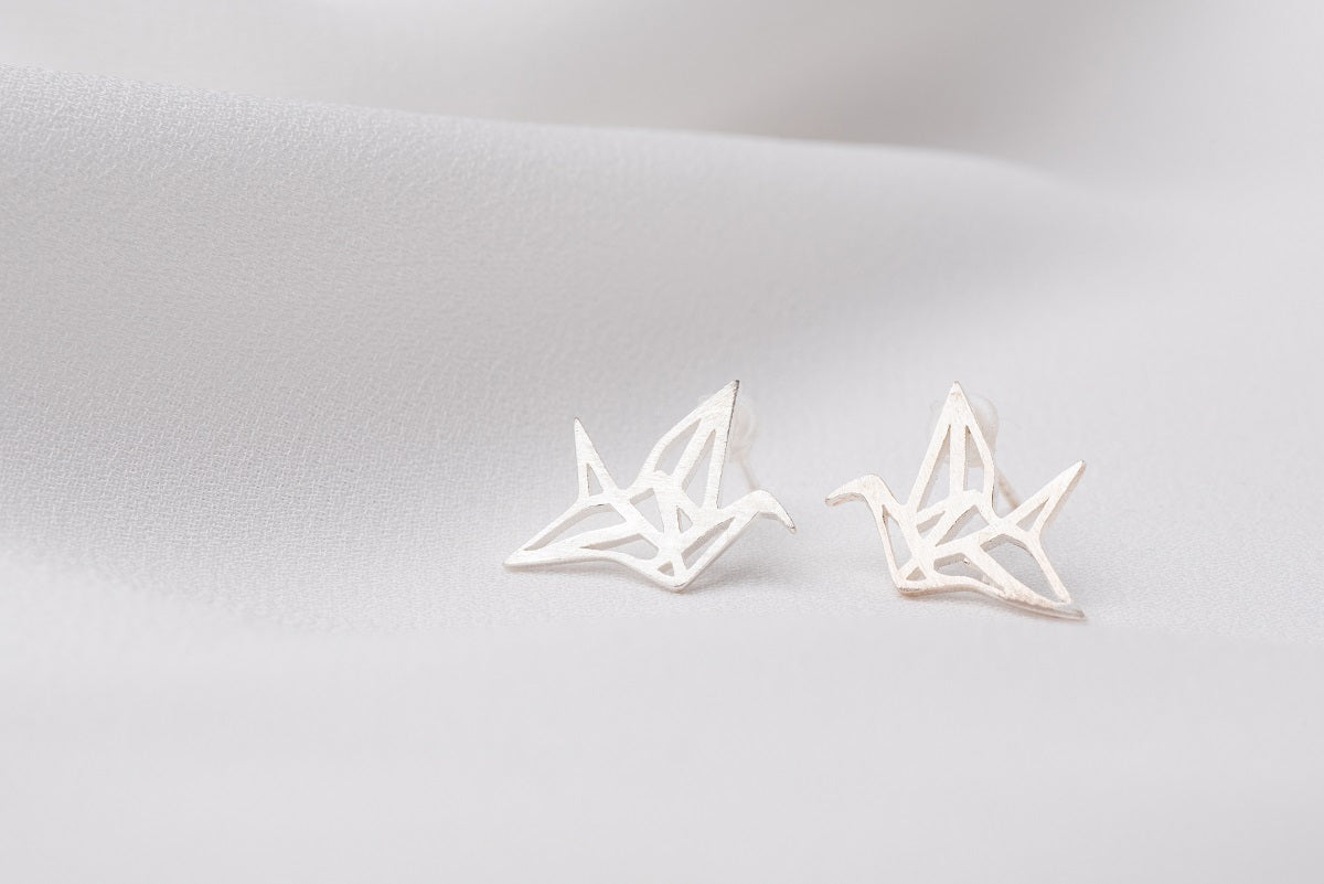 A pair of silver stud earrings designed in a origami crane shape, placed on a white surface.