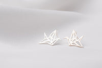 A pair of silver stud earrings designed in a origami crane shape, placed on a white surface.