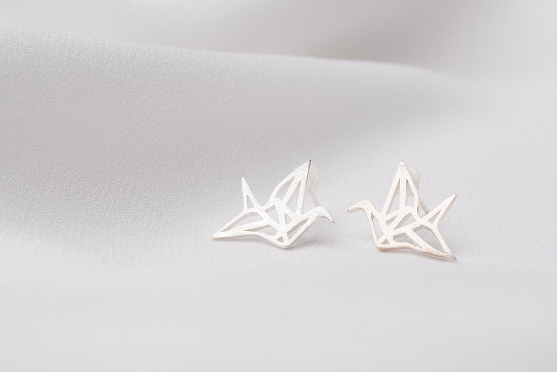 A pair of silver stud earrings designed in a origami crane shape, placed on a white surface.