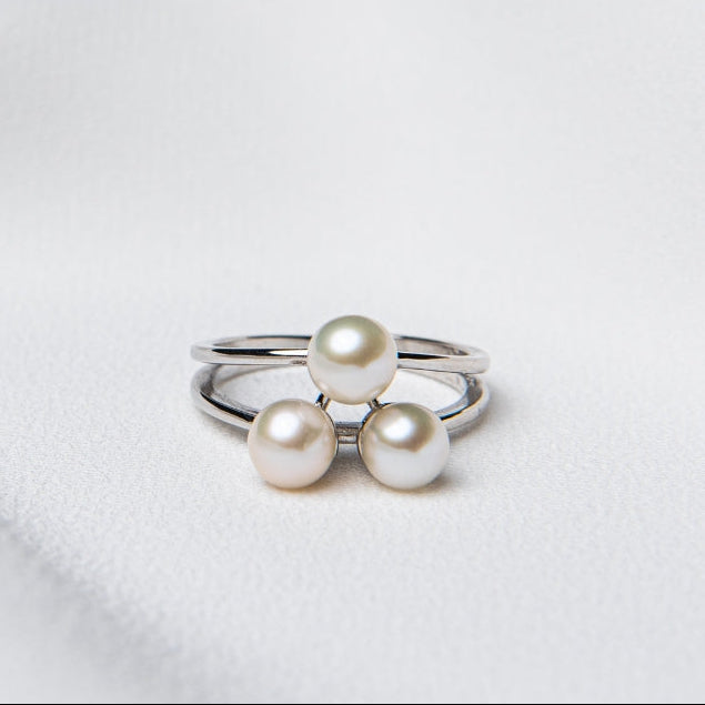 An adjustable size ring with a polished finish, featuring a silver band and three round white pearls on top, placed on a white fabric surface.