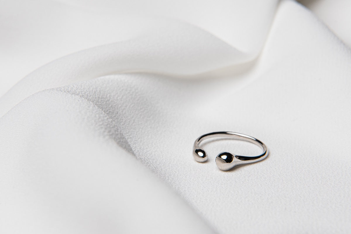 A polished, silver-colored ring with a simple design, displayed on a white fabric surface.
