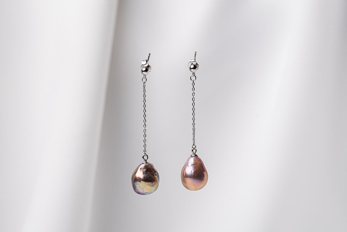 A pair of baroque style pearl earrings with silver chains and butterfly back closures, displayed against a white fabric background.