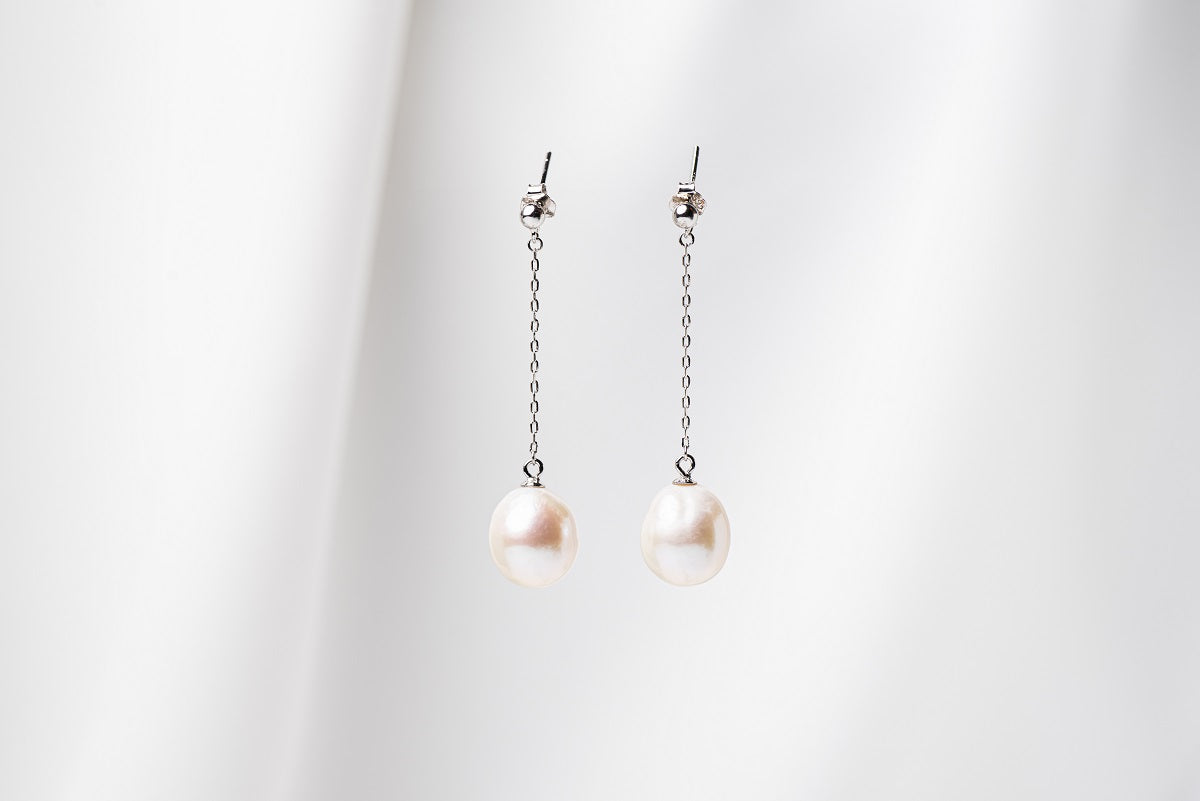 A pair of baroque style pearl earrings with silver chains and butterfly back closures, displayed against a white fabric background.