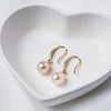 A pair of dangle champaigne pearl hook earrings in gold vermeil, placed inside a white heart-shaped bowl on a white surface.
