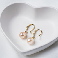 A pair of dangle champaigne pearl hook earrings in gold vermeil, placed inside a white heart-shaped bowl on a white surface.