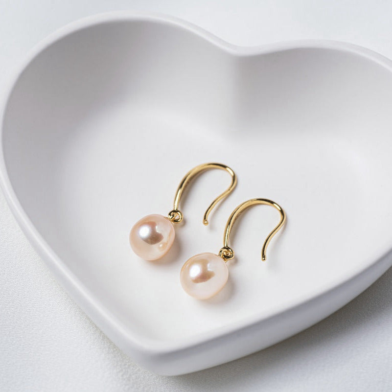 A pair of dangle champaigne pearl hook earrings in gold vermeil, placed inside a white heart-shaped bowl on a white surface.