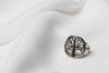 A silver ring with a tree of life design, displayed on a white textured surface.
