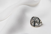 A silver ring with a tree of life design, displayed on a white textured surface.