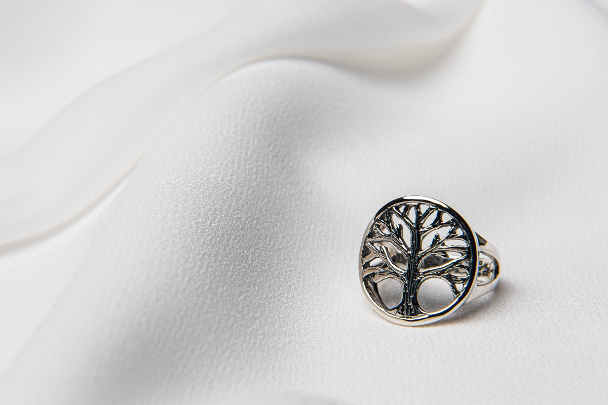 A silver ring with a tree of life design, displayed on a white textured surface.